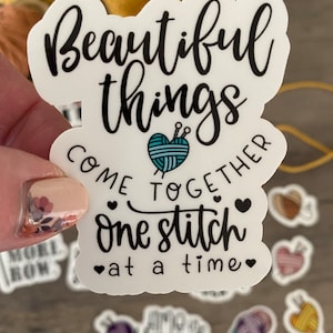 May include: A white sticker with black script that reads "Beautiful things come together one stitch at a time." A teal yarn ball with needles is in the center. Small heart graphics are included.