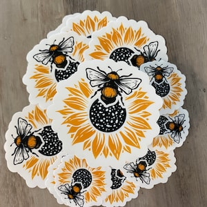 May include: A stack of round stickers featuring a black and yellow bumblebee on a yellow sunflower with a black center. The stickers are white with a scalloped edge.
