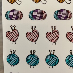 May include: A sheet of stickers featuring various yarn balls and hearts with knitting needles. The stickers are in shades of pink, blue, purple, and brown.