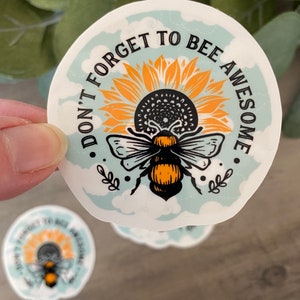 May include: A round sticker with a blue background and white clouds. The sticker features a bee with yellow and black stripes, sitting on a sunflower with yellow petals and a brown center. The text "Don't forget to bee awesome" is written in black around the bee and sunflower.