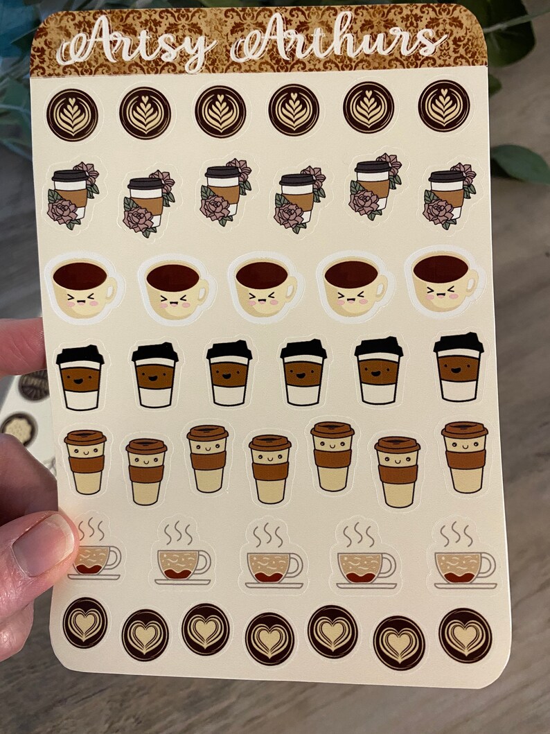 Pu&ograve; includere: Un foglio di adesivi con vari disegni di tazze da caff&egrave;. Gli adesivi sono su uno sfondo beige con un bordo marrone. I disegni includono tazze da caff&egrave; con diverse espressioni facciali, tazze da caff&egrave; con fiori e tazze da caff&egrave; con vapore che esce da esse. Il testo "Artsy Arthurs" &egrave; in cima al foglio.
