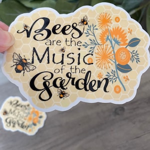 May include: A yellow and white sticker with a honeycomb pattern and floral designs. The sticker says "Bees are the Music of the Garden".
