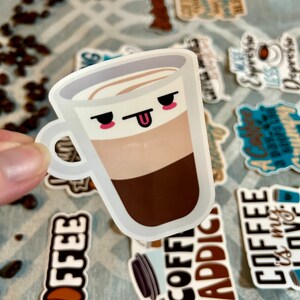 以下が含まれることがあります： 顔と突き出た舌のあるコーヒーカップの漫画のイラストが描かれた白いステッカー。カップは、茶色のコーヒー層、ベージュのミルク層、白い泡層を持っています。ステッカーは、他のコーヒーをテーマにしたステッカーのあるグレーとホワイトのパターンで覆われた表面にあります。