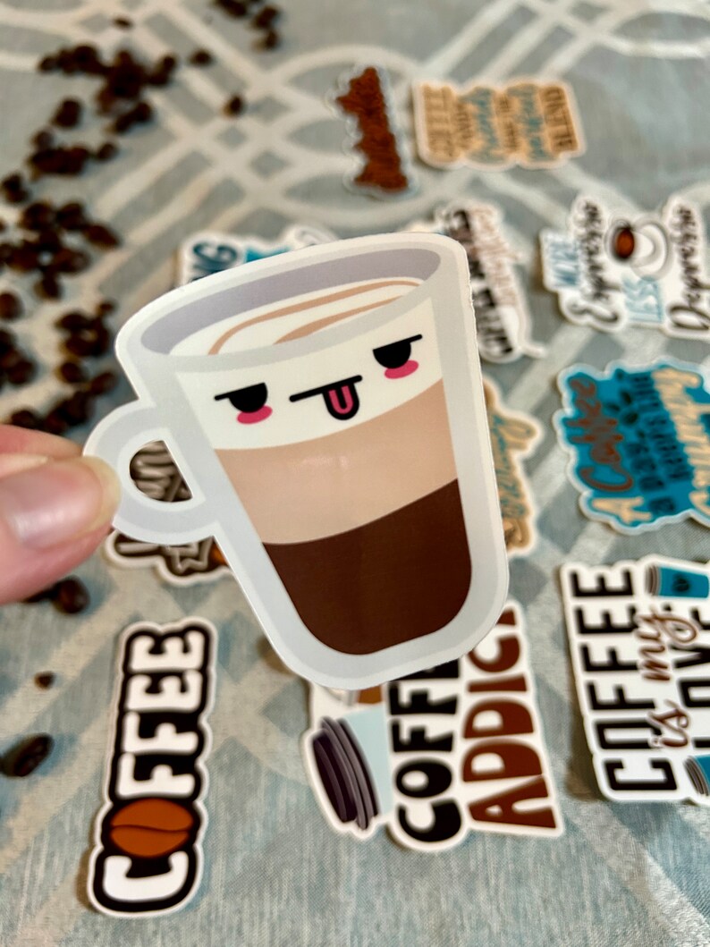 以下が含まれることがあります： 顔と突き出た舌のあるコーヒーカップの漫画のイラストが描かれた白いステッカー。カップは、茶色のコーヒー層、ベージュのミルク層、白い泡層を持っています。ステッカーは、他のコーヒーをテーマにしたステッカーのあるグレーとホワイトのパターンで覆われた表面にあります。