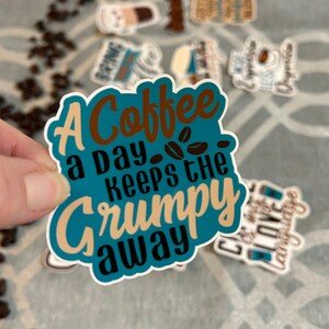 以下が含まれることがあります： 茶色、黒、ベージュの文字で「A Coffee a Day Keeps the Grumpy Away」と書かれたターコイズブルーのステッカー。ステッカーにはコーヒー豆のデザインが施され、他のコーヒーをテーマにしたステッカーに囲まれています。
