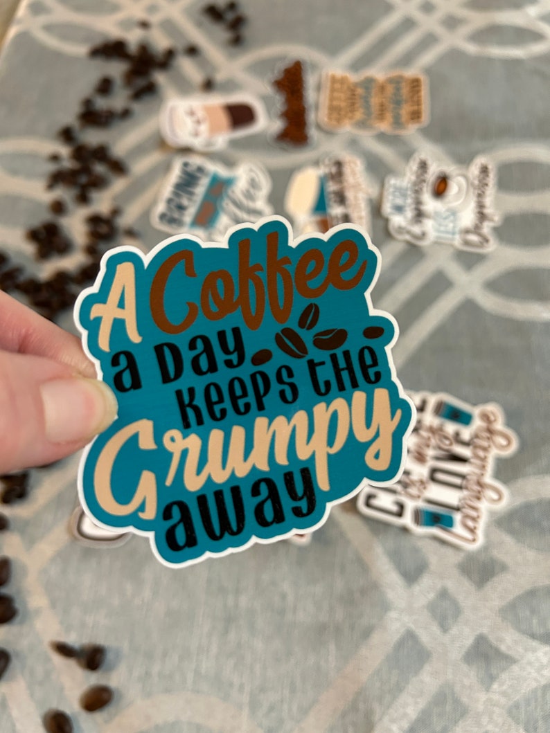 以下が含まれることがあります： 茶色、黒、ベージュの文字で「A Coffee a Day Keeps the Grumpy Away」と書かれたターコイズブルーのステッカー。ステッカーにはコーヒー豆のデザインが施され、他のコーヒーをテーマにしたステッカーに囲まれています。