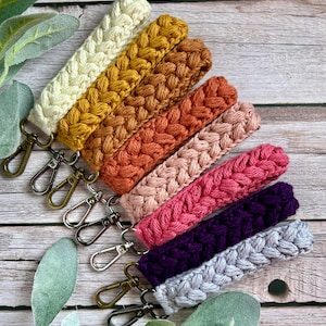 May include: A collection of handmade crochet wristlets in various colors, including cream, mustard yellow, orange, pink, purple, and gray. Each wristlet features a braided design and a metal clasp for attaching keys or other items.
