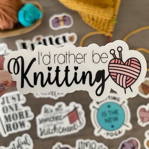 May include: A white sticker with the text "I'd rather be Knitting" in black script. A pink yarn heart with knitting needles is to the right of the text. Other stickers with knitting-related phrases and images are scattered around.