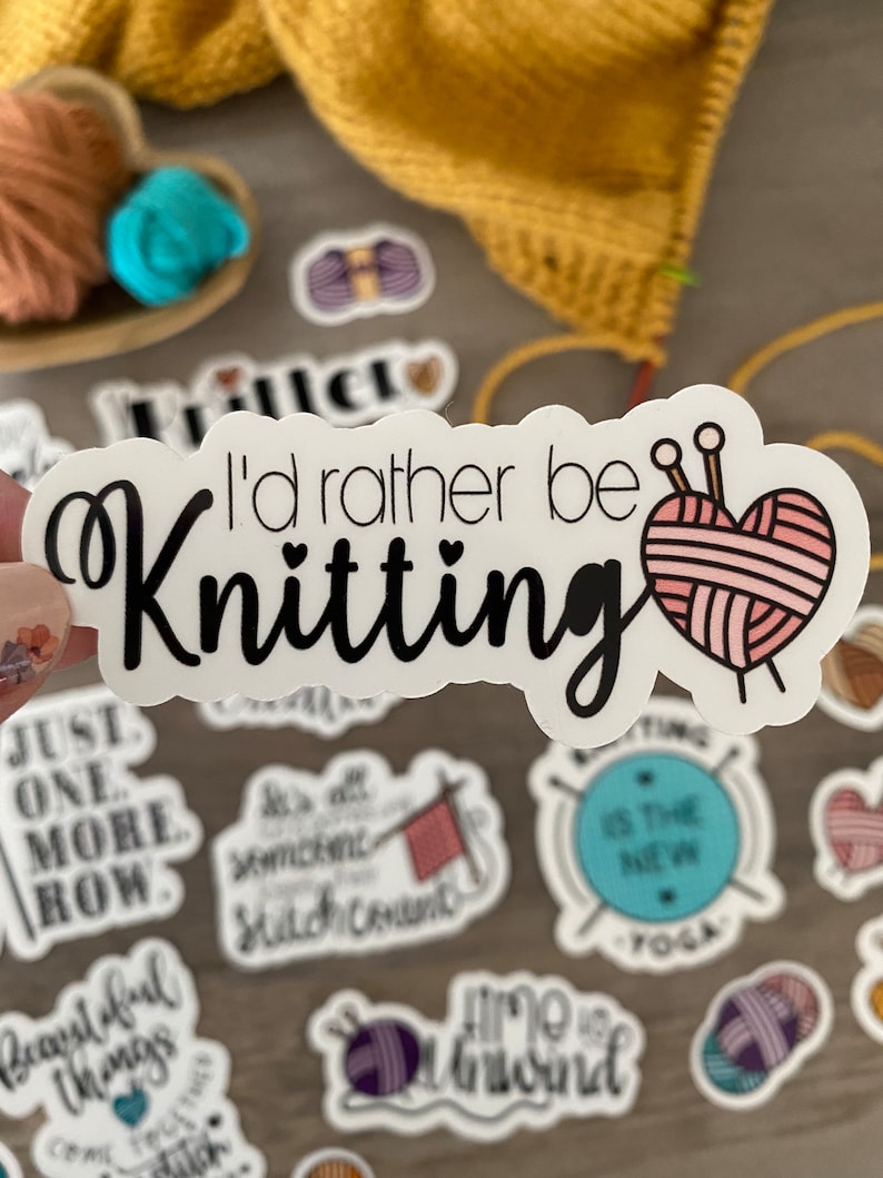 May include: A white sticker with the text "I'd rather be Knitting" in black script. A pink yarn heart with knitting needles is to the right of the text. Other stickers with knitting-related phrases and images are scattered around.