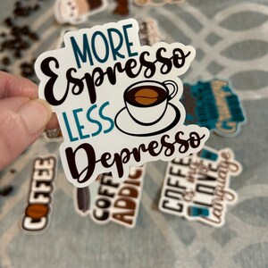 以下が含まれることがあります： 黒と青の文字で「More Espresso Less Depresso」と書かれたステッカー。ステッカーの中央には、茶色のコーヒー豆が入った小さな白いカップが描かれています。