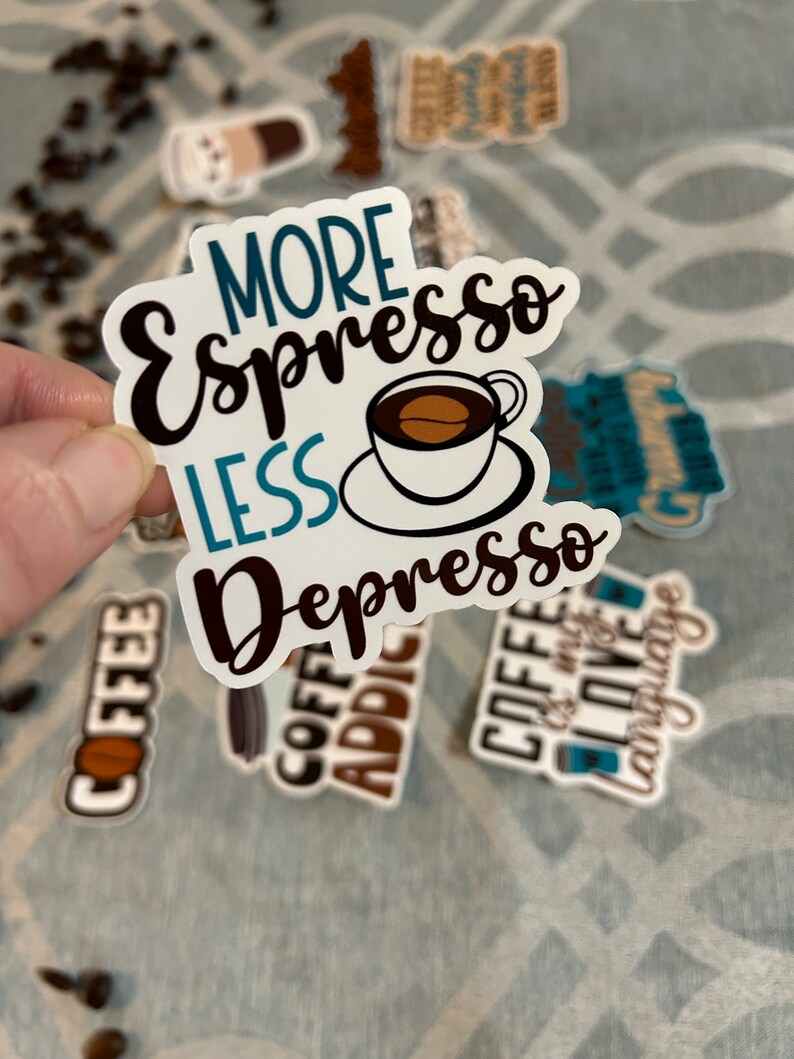 以下が含まれることがあります： 黒と青の文字で「More Espresso Less Depresso」と書かれたステッカー。ステッカーの中央には、茶色のコーヒー豆が入った小さな白いカップが描かれています。