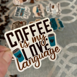 以下が含まれることがあります： 白地に茶色と黒の文字で「Coffee is my love language」と書かれたステッカー。ステッカーの右側に、コーヒー豆が描かれた青いコーヒーカップがある。