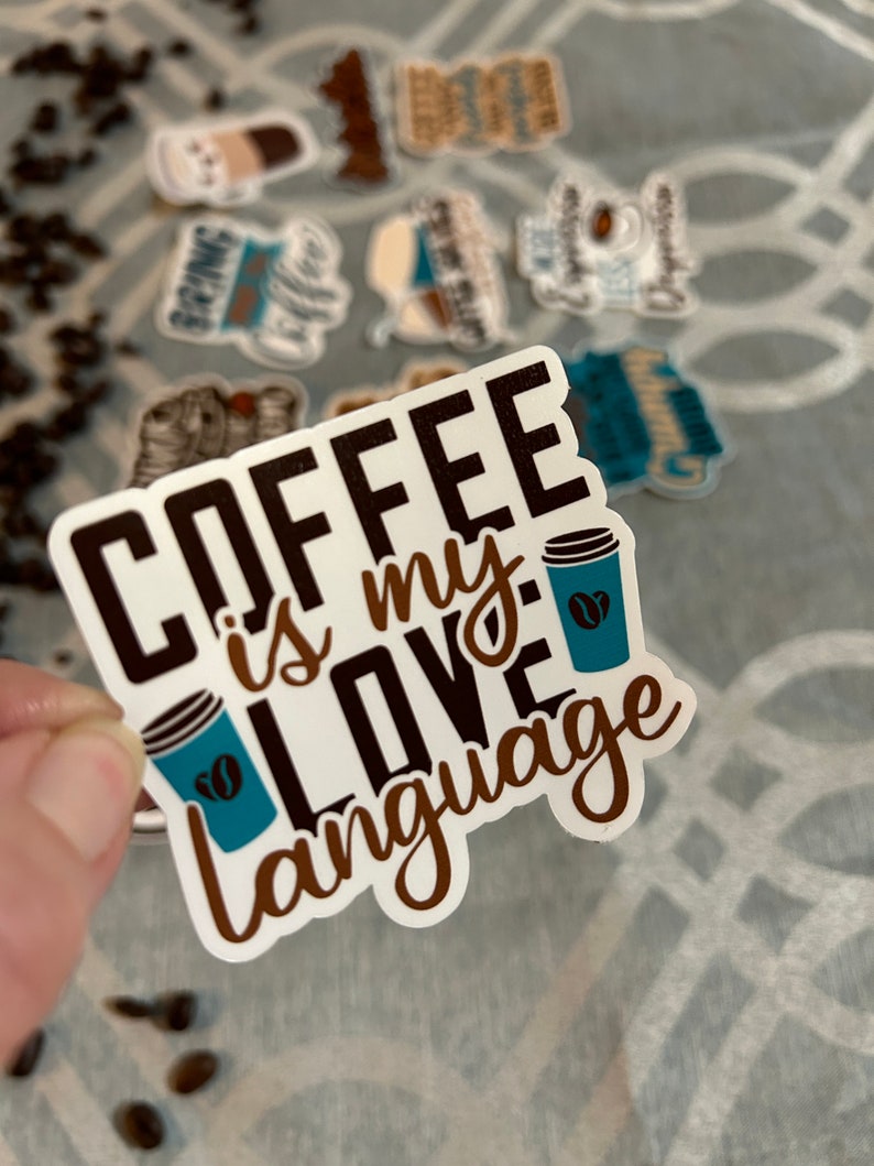 以下が含まれることがあります： 白地に茶色と黒の文字で「Coffee is my love language」と書かれたステッカー。ステッカーの右側に、コーヒー豆が描かれた青いコーヒーカップがある。
