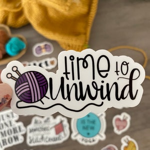 May include: A white sticker with the words "time to unwind" in black script. The sticker features a purple yarn ball with knitting needles. Other stickers with knitting themes are also visible.