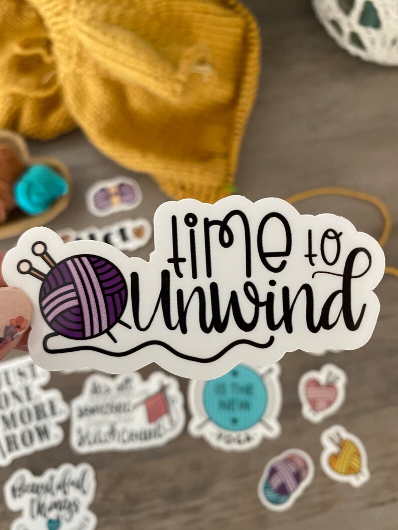 May include: A white sticker with the words "time to unwind" in black script. The sticker features a purple yarn ball with knitting needles. Other stickers with knitting themes are also visible.