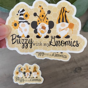 May include: Three gnomes with bee wings and black and yellow striped hats are on a yellow honeycomb background. The text "Buzzing with my Nomies" is below the gnomes.