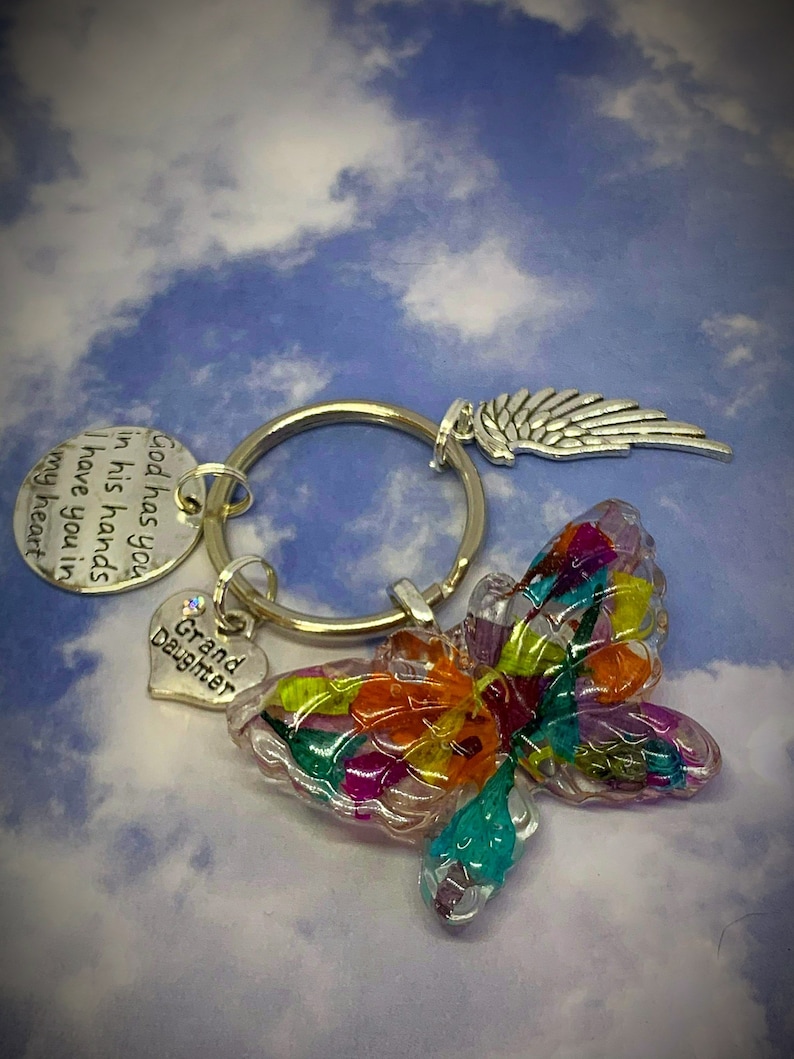 May include: A silver keychain with a colorful butterfly charm, a heart charm that says "Grand Daughter", and a wing charm. The keychain also has a silver tag with the text "God has you in his hands I have you in my heart."