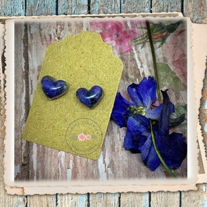 May include: A pair of heart-shaped resin earrings with pressed purple flowers inside. The earrings are displayed on a brown card with the text "Simply Gorgeous" and a floral design.