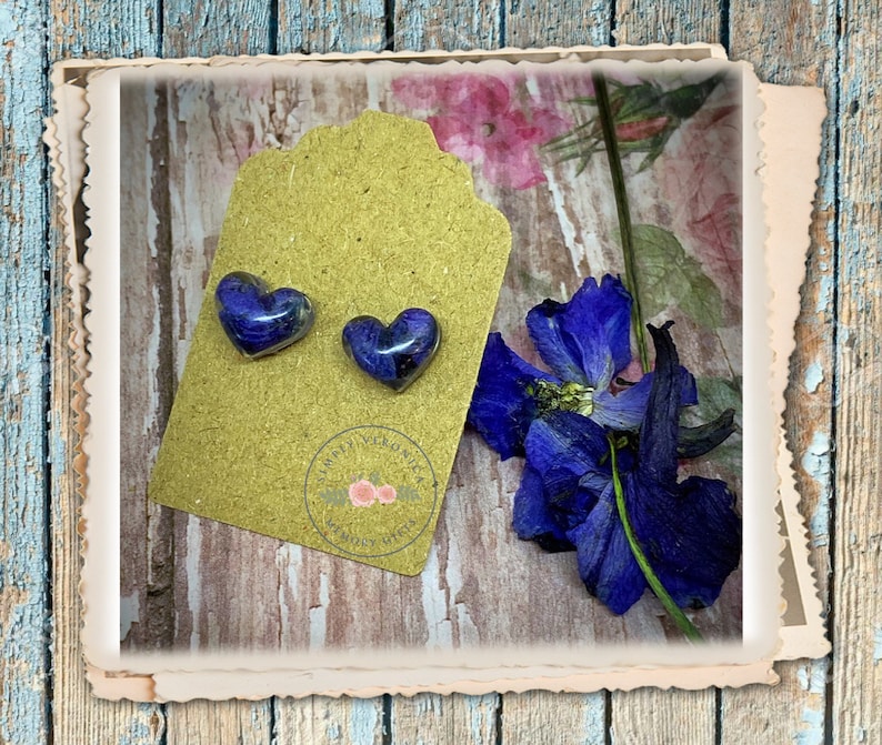 May include: A pair of heart-shaped resin earrings with pressed purple flowers inside. The earrings are displayed on a brown card with the text "Simply Gorgeous" and a floral design.