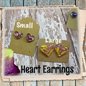 May include: Two pairs of heart-shaped earrings with pressed flowers inside. The smaller pair has pink flowers and the larger pair has yellow and pink flowers. The earrings are displayed on brown cards with the words "Small" and "Large" printed on them. The text "Heart Earrings" is printed below the earrings.