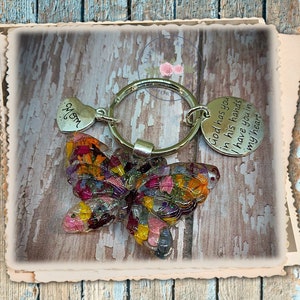 May include: A silver key chain with a butterfly charm made of pressed flowers. The butterfly is multi-colored with shades of pink, yellow, orange, and purple. The key chain also has a heart charm with the word "Mom" engraved on it and a round charm with the text "God has you in his hands I have you in my heart".