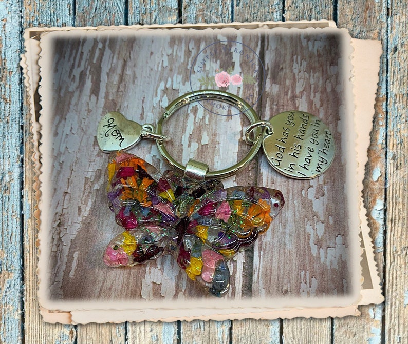 May include: A silver key chain with a butterfly charm made of pressed flowers. The butterfly is multi-colored with shades of pink, yellow, orange, and purple. The key chain also has a heart charm with the word "Mom" engraved on it and a round charm with the text "God has you in his hands I have you in my heart".