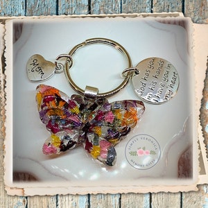 May include: A silver key chain with a butterfly charm made of pressed flowers. The butterfly is multi-colored with shades of pink, yellow, orange, and purple. The key chain also has a heart charm with the word "Mom" on it and a round charm with the text "God has you in his hands have you in my heart". The key chain is from Simply Veronica Memory Gifts.