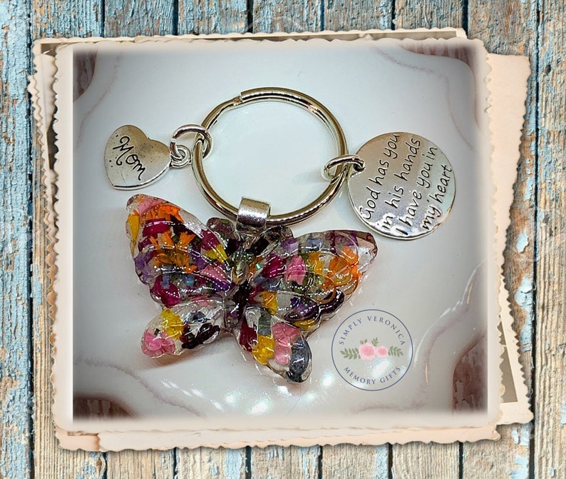 May include: A silver key chain with a butterfly charm made of pressed flowers. The butterfly is multi-colored with shades of pink, yellow, orange, and purple. The key chain also has a heart charm with the word "Mom" on it and a round charm with the text "God has you in his hands have you in my heart". The key chain is from Simply Veronica Memory Gifts.