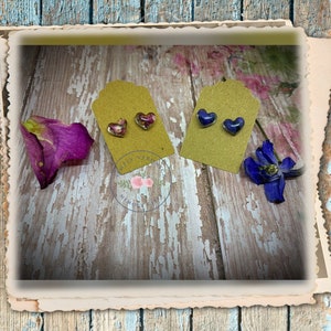 May include: Two pairs of heart-shaped resin earrings with pressed flowers inside. One pair has pink and purple flowers, the other has blue flowers.