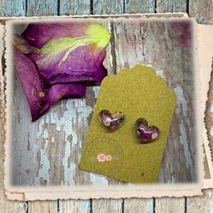 May include: A pair of heart-shaped earrings with dried pink and purple flowers encased in clear resin. The earrings are displayed on a brown tag with a gold glitter finish.