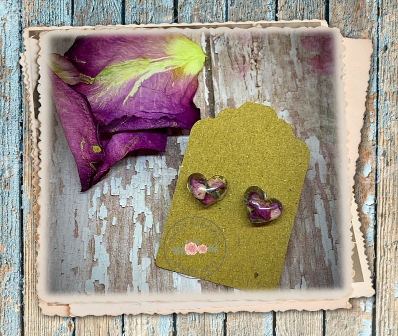 May include: A pair of heart-shaped earrings with dried pink and purple flowers encased in clear resin. The earrings are displayed on a brown tag with a gold glitter finish.