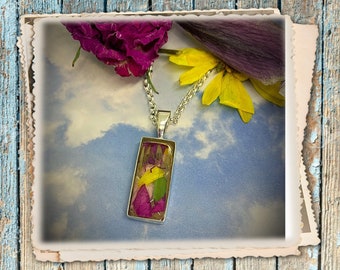 Op de afbeelding: Een zilveren ketting met een rechthoekige hanger met geperste bloemen in roze, gele en groene tinten. De hanger is geplaatst tegen een bewolkte blauwe achtergrond.