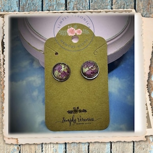 May include: A pair of silver stud earrings with a round, clear resin setting. The resin contains pressed purple and white flowers. The earrings are displayed on a brown card with the text "Simply Veronica Memory Gifts".