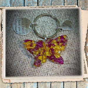 May include: A butterfly-shaped keychain with a resin-filled design featuring yellow and pink flowers. The keychain includes a silver ring and two charms: one with the text "God has you in his hands I have you in my heart" and the other with the word "Grandma".