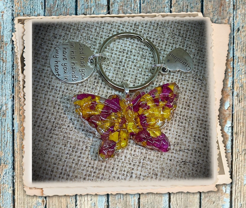 May include: A butterfly-shaped keychain with a resin-filled design featuring yellow and pink flowers. The keychain includes a silver ring and two charms: one with the text "God has you in his hands I have you in my heart" and the other with the word "Grandma".