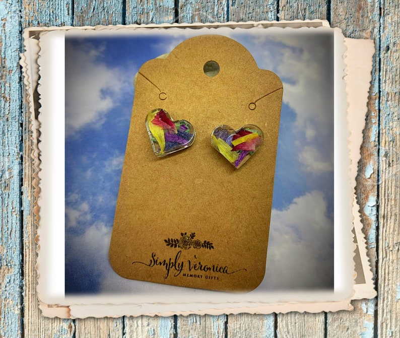 May include: A pair of heart-shaped earrings with pressed flowers encased in clear resin. The earrings are on a brown card with the text "Simply Veronica Memory Gifts".