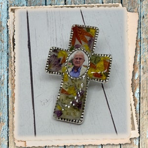 May include: A handcrafted cross pendant with a silver-colored border. The cross is filled with dried flowers and a small oval photo of an elderly person. The background is a light blue and white wooden surface.