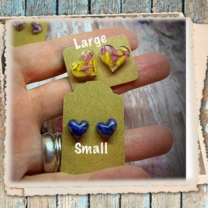 May include: Two pairs of heart-shaped earrings. The larger pair is yellow and pink with dried flowers encased in resin. The smaller pair is blue with a darker blue swirl in the resin. The earrings are displayed on a brown card with the words "Large" and "Small" printed on them.