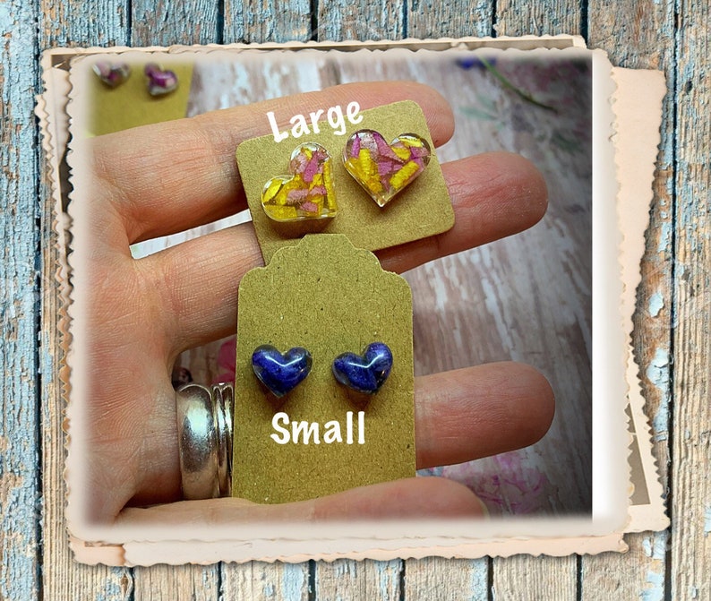 May include: Two pairs of heart-shaped earrings. The larger pair is yellow and pink with dried flowers encased in resin. The smaller pair is blue with a darker blue swirl in the resin. The earrings are displayed on a brown card with the words "Large" and "Small" printed on them.