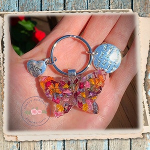 May include: A silver butterfly keychain with pressed flowers in shades of pink, orange, and yellow. The keychain has a silver heart charm with the word "Mom" and a round charm with the text "God has you in his hands I have your heart".