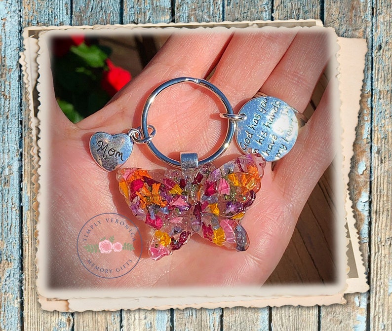 May include: A silver butterfly keychain with pressed flowers in shades of pink, orange, and yellow. The keychain has a silver heart charm with the word "Mom" and a round charm with the text "God has you in his hands I have your heart".