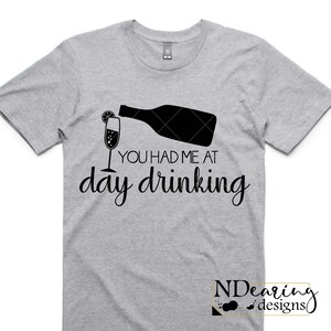 May include: A light gray t-shirt with a black graphic of a champagne bottle pouring into a glass with a lemon slice. The text "YOU HAD ME AT day drinking" is below the graphic. The shirt has short sleeves.