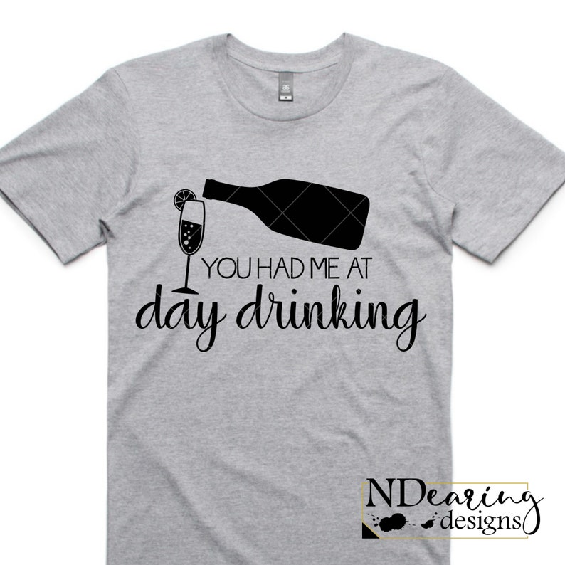 May include: A light gray t-shirt with a black graphic of a champagne bottle pouring into a glass with a lemon slice. The text "YOU HAD ME AT day drinking" is below the graphic. The shirt has short sleeves.