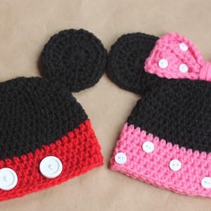 May include: Two crocheted hats with Mickey Mouse ears. One hat is black with red trim and white buttons. The other hat is black with pink trim and white buttons. Both hats have a bow on top.