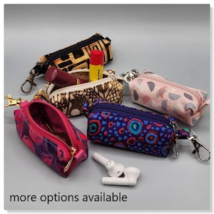 Puede incluir: Cinco pequeñas bolsas con cremallera y llaveros, cada una con una tela estampada de colores diferente. Las bolsas están dispuestas sobre una superficie gris con un par de auriculares inalámbricos blancos.