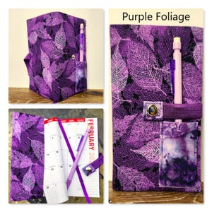 Op de afbeelding: Een stoffen plannerhoes met een paars bladskeletpatroon op een zwarte achtergrond. De hoes bevat een penlus, een klein zakje en een drukknoopsluiting. De tekst Purple Foliage is zichtbaar. Een kalender is in de hoes geplaatst.