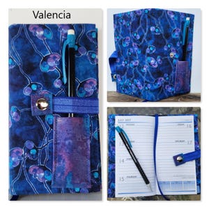 Op de afbeelding: Een stoffen plannerhoes met een donkerblauwe achtergrond en een patroon van paarse en blauwgroene abstracte bloemen. De hoes bevat een elastische band met een metalen drukknoop, een penhouder en een zwarte pen. De tekst Valencia is zichtbaar.