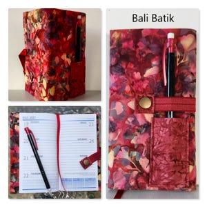 Op de afbeelding: Een stoffen notitieboekhoes met een rood, roze en geel Bali Batik bloemenpatroon. De hoes heeft een buitenvak, een penlus met een zwarte pen en een drukknoopsluiting. De binnenzijde bevat een 2027-planner met een rood lint als bladwijzer.