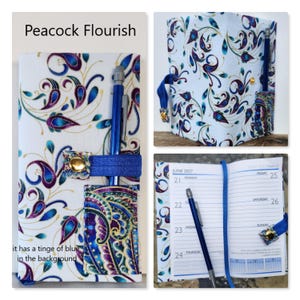 Op de afbeelding: Een stoffen plannerhoes met de titel Peacock Flourish, voorzien van een witte achtergrond met blauwe, paarse en gouden paisley-patronen. Het bevat een blauwe elastische band met een gouden drukknoop, een pennenhouder en een open planner met lijntjes.