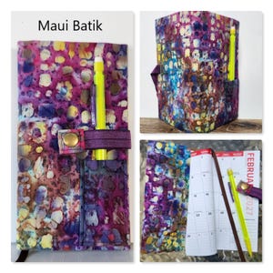 Op de afbeelding: Een stoffen plannerhoes met een Maui Batik-patroon in paarse, roze, blauwe en gele tinten. De hoes is voorzien van een elastische band met een koperen drukknoopsluiting en een klein vakje waarin een felgele pen is opgeborgen.